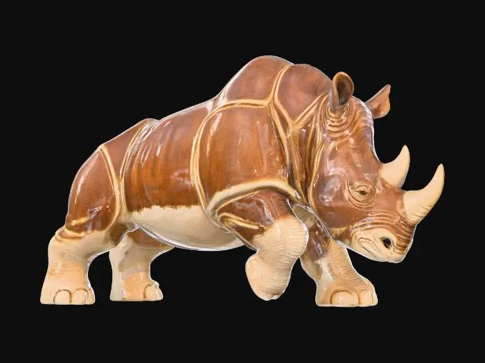 3D model for A powerful ceramic rhinoceros figurine, depicted mid-charge with its head lowered and short, thick legs pushing forward. Its robust, chunky body is coated in a glossy two-tone glaze: a dark, earthy brown for its armor-like skin, highlighted by creamy beige on its underbelly and inner limbs. The sculpted form is smooth and cool, reflecting light intensely, conveying raw, unstoppable force.