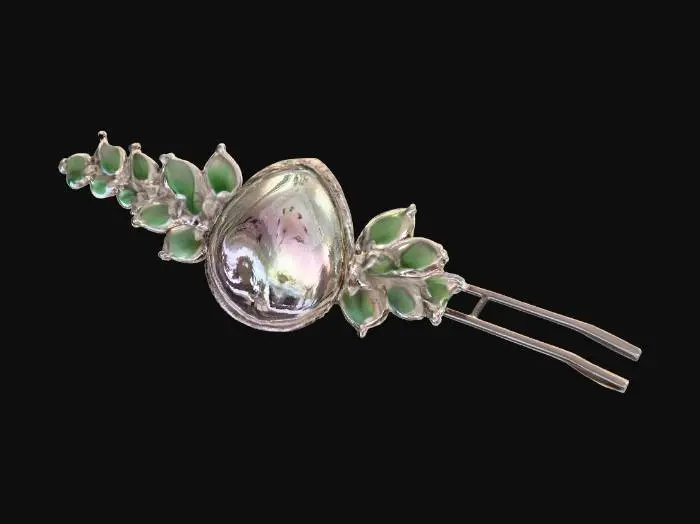3D model for A long, slender hair pin, characteristic of Victorian elegance, features a silver body intricately embossed with floral motifs. The head of the pin is adorned with a large, teardrop-shaped mother of pearl piece, exhibiting a creamy white color with faint pink and green overtones. It is framed by a delicate border of minute, square-cut emeralds and tiny, round sapphires, creating a captivating sparkle.