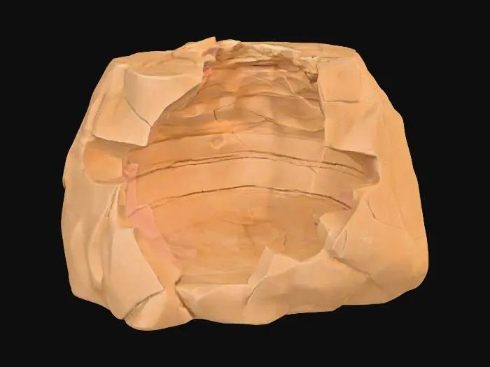 3D model for A broken sandstone rib segment, presenting a distinct missing chunk on one end, revealing the raw, fractured interior of the rock. The remaining edge around this void features a jagged, fractured lip, indicating natural breakage and erosion. Despite the damage, its layered sedimentary strata maintain a consistent direction, suggesting its origin from a larger, continuous formation. Composed of granular sandstone, it exhibits warm ochre and reddish-brown tones, with a rough, weathered texture across its fragmented form.