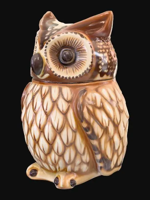 3D model for A charming, small trinket jar is meticulously crafted to resemble a plump owl. Made from smooth, glazed ceramic, its surface is cool and hard to the touch, painted in warm, earthy tones of brown and cream, with distinct feathery patterns. The owl's head is a separate piece, serving as the lid, designed to fit snugly onto the hollow body, creating a seamless appearance when closed. Its large, round eyes give it a wise, whimsical expression.