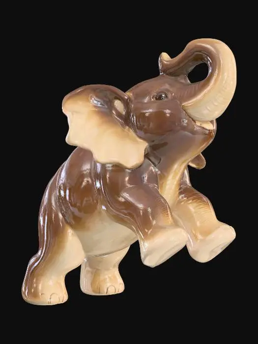 3D model for A playful ceramic elephant calf figurine, depicted with its trunk playfully raised and ears flapping, in a short, robust stance. Its rotund body and thick limbs are covered in a glossy two-tone glaze: a rich, dark brown on its back and outer legs, complemented by a soft, creamy beige on its belly and ear interiors. The smooth, cool sculpture feels solid and substantial, conveying youthful exuberance.
